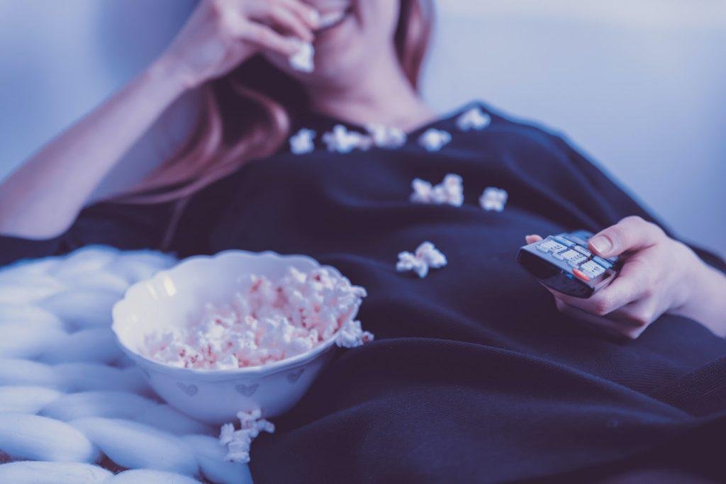 woman lying on bed while eating puff corn and watching movie