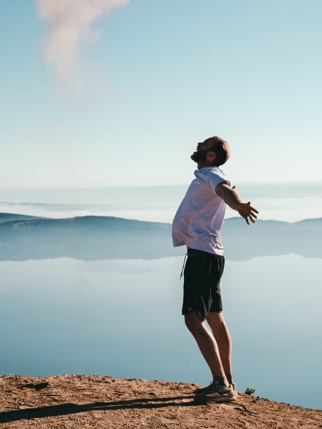 man standing on sand while spreading arms beside calm body of water